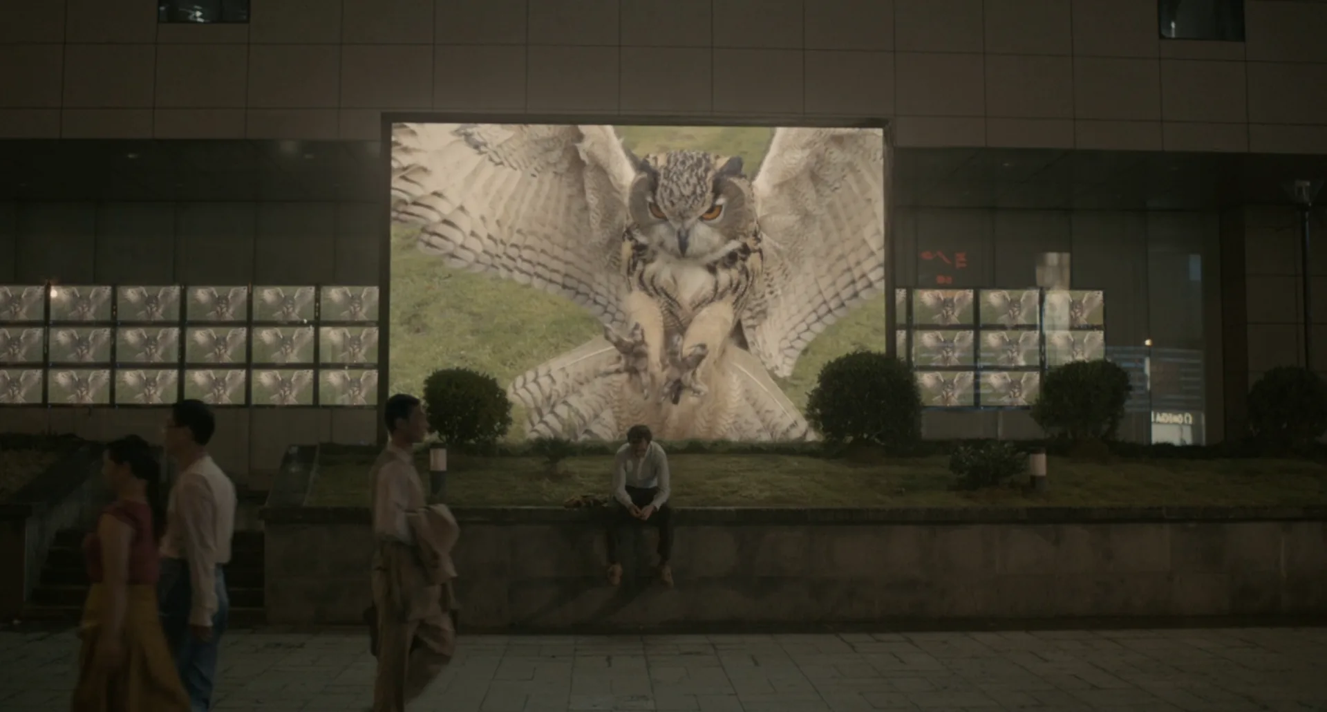 Theodore sentado, pequeño, frente a una pantalla pública gigante en la ciudad, en la película Her