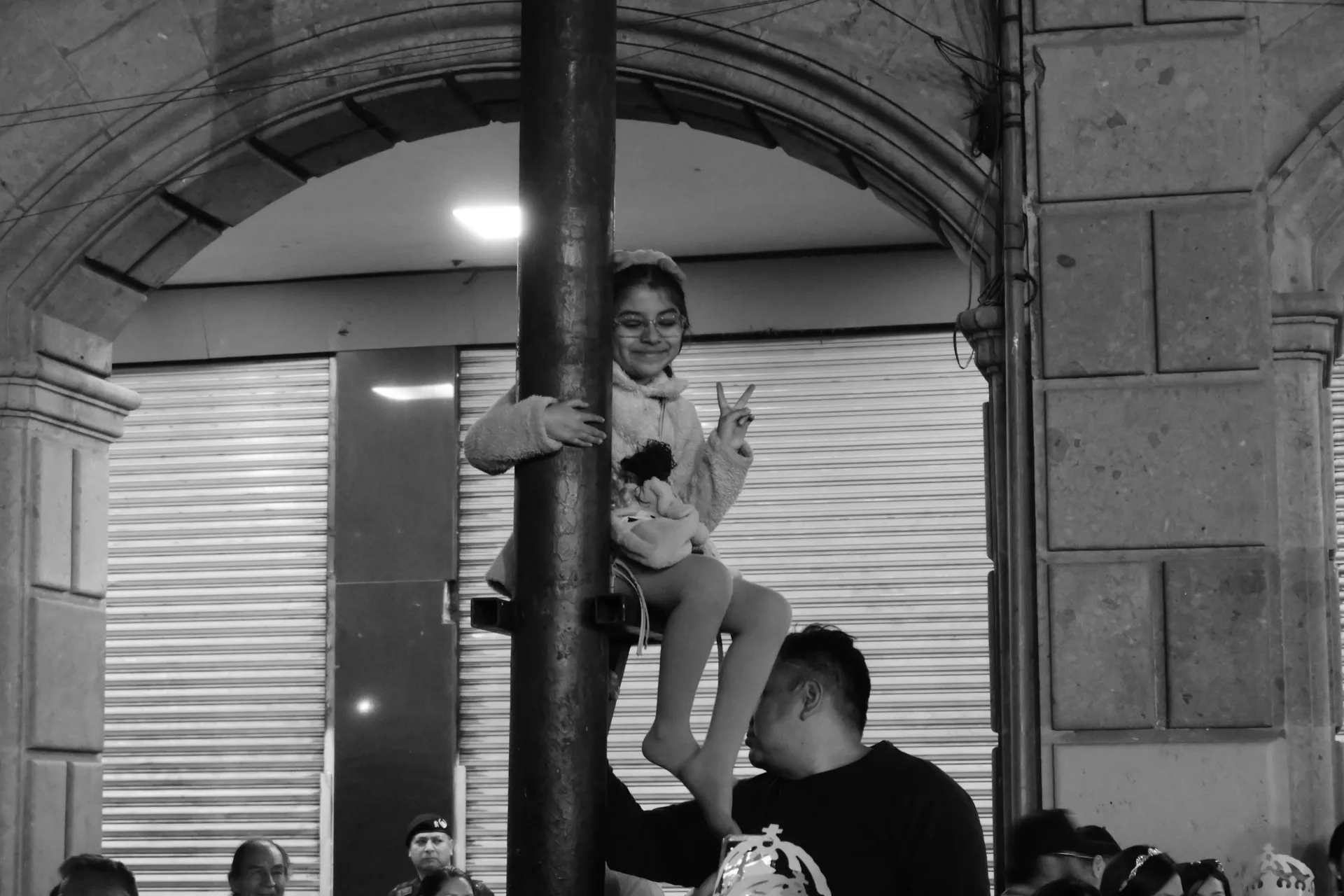 Niña subida a un poste sostenida por un hombre, haciendo señal de paz entre la multitud