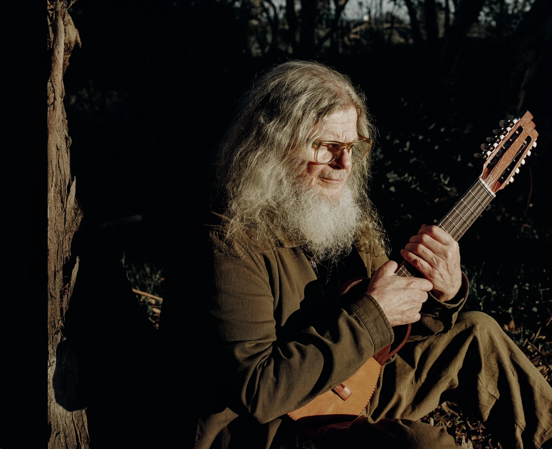 Santaolalla recargado contra un árbol al atardecer, tocando un charango con los ojos cerrados, luz dorada lateral
