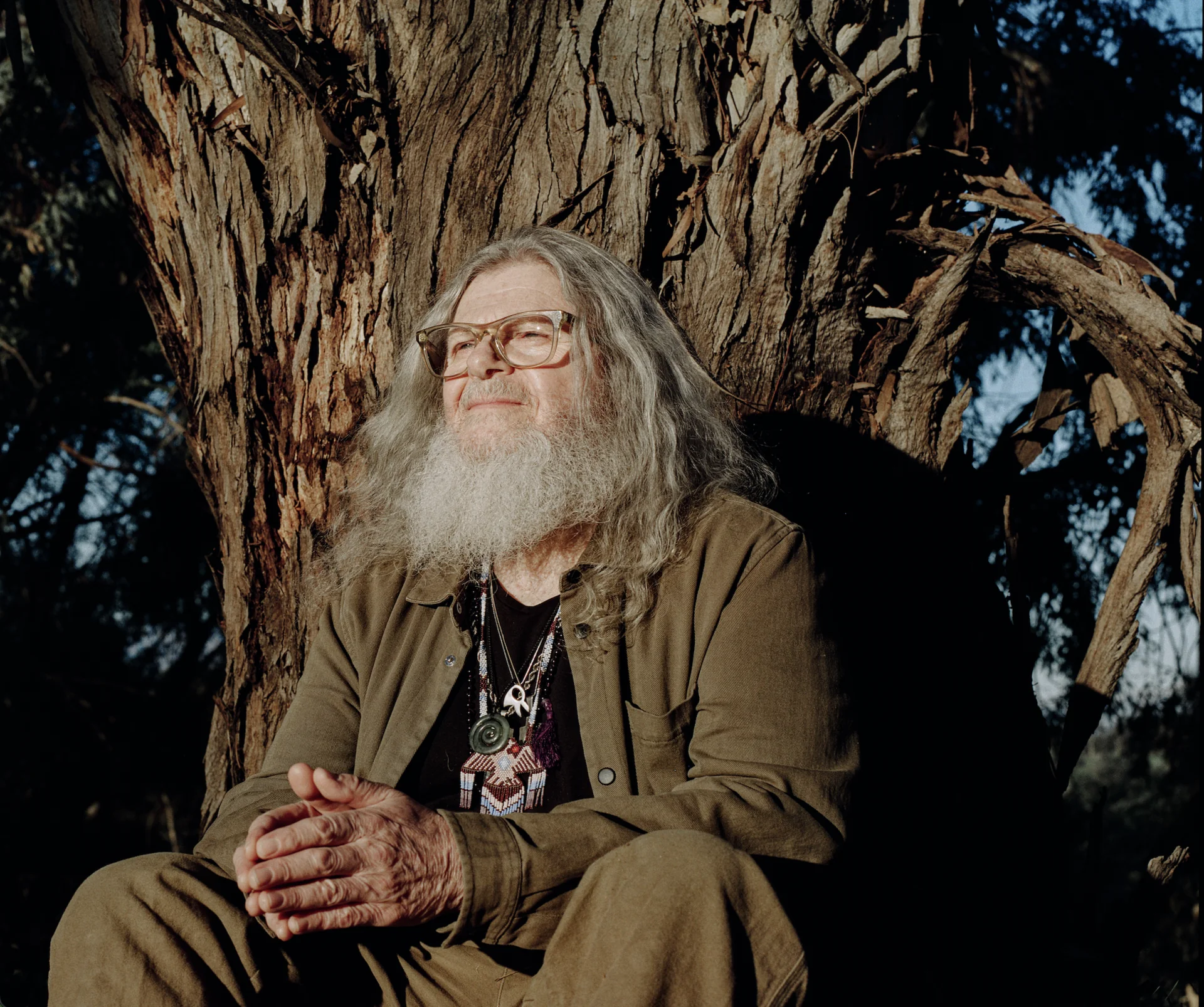 Santaolalla sentado contra el tronco de un árbol, mirando hacia arriba con una sonrisa amplia, luz natural
