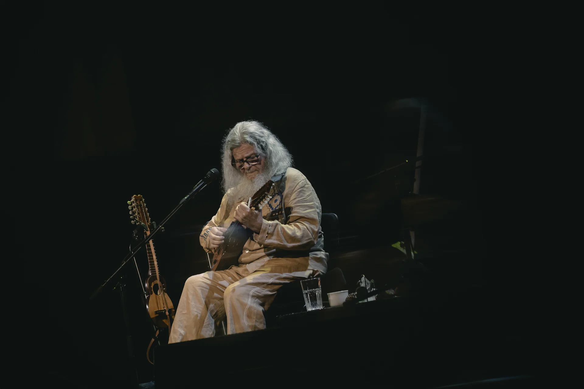Santaolalla solo en escenario tocando un instrumento de cuerda, ronroco apoyado a su lado, luz puntual sobre el músico, fondo negro