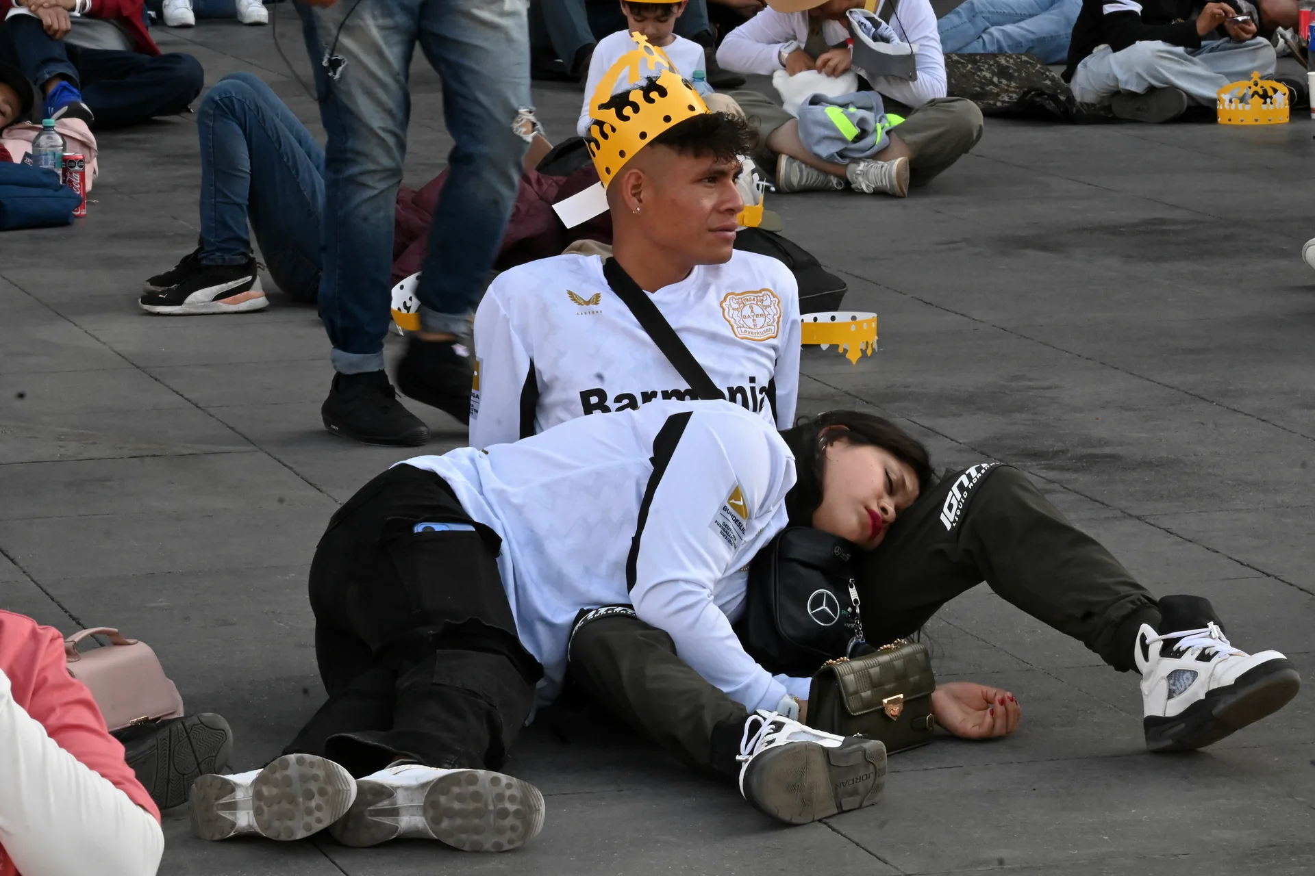 Pareja en el suelo del Zócalo: él con corona de papel de la marca Corona en la cabeza, ella dormida sobre sus piernas durante la espera previa al concierto