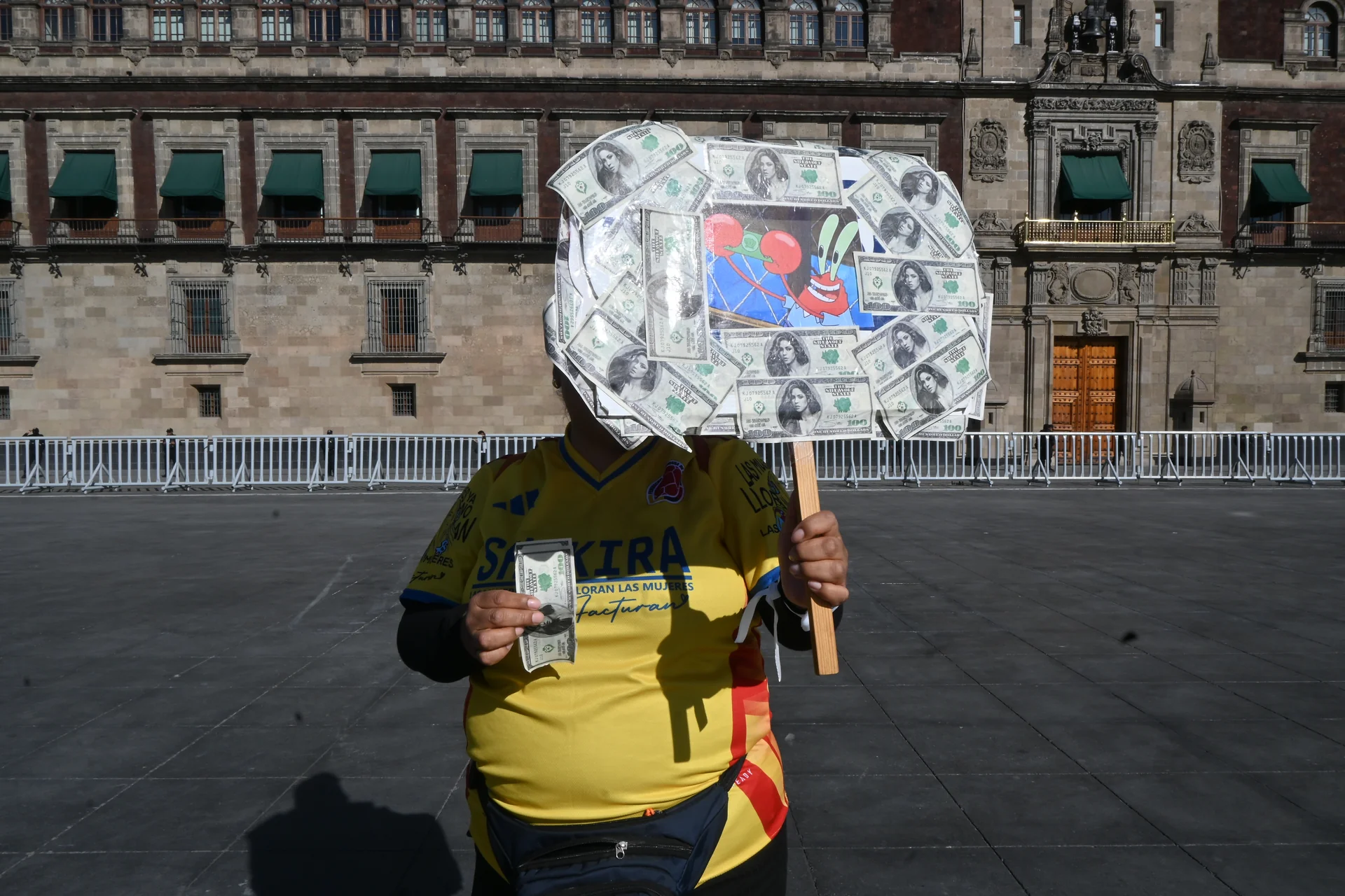 Vendedora ambulante con jersey del América sostiene un cartel con billetes de un dólar con la cara de Shakira a diez pesos cada uno, en el Zócalo de día